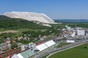 Industriegelände mit weißen Lagerhügeln, Fabrikgebäuden, Bahnschienen und Waggons, umgeben von bewaldeten Hügeln.