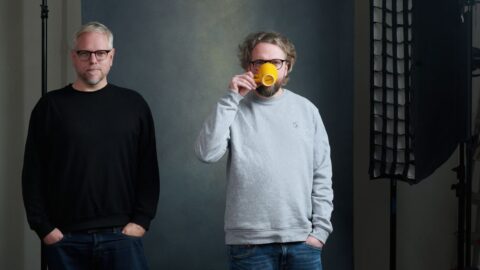 Zwei Männer stehen nebeneinander, einer hält eine gelbe Tasse, vor einem neutralen Hintergrund mit Studiobeleuchtung.