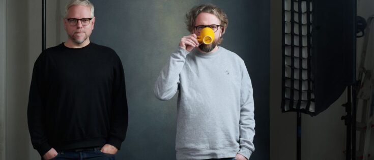 Zwei Männer stehen nebeneinander, einer hält eine gelbe Tasse, vor einem neutralen Hintergrund mit Studiobeleuchtung.