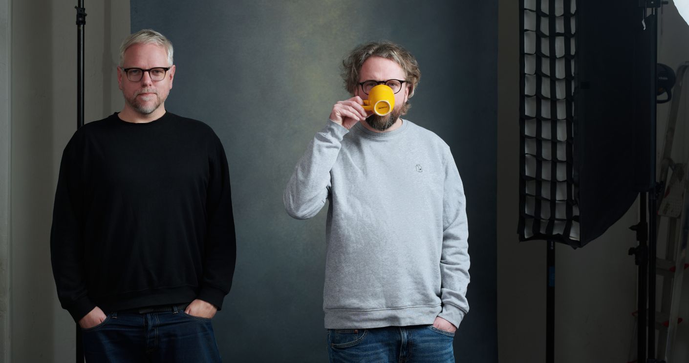 Zwei Männer stehen nebeneinander, einer hält eine gelbe Tasse, vor einem neutralen Hintergrund mit Studiobeleuchtung.