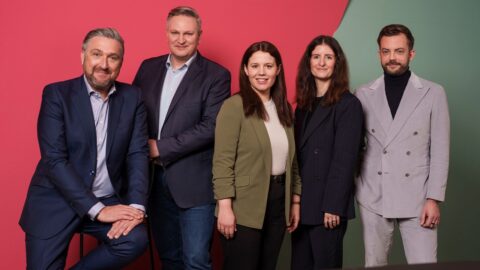 Fünf Personen in formeller Kleidung stehen vor einem bunten Hintergrund aus Pink und Grün.