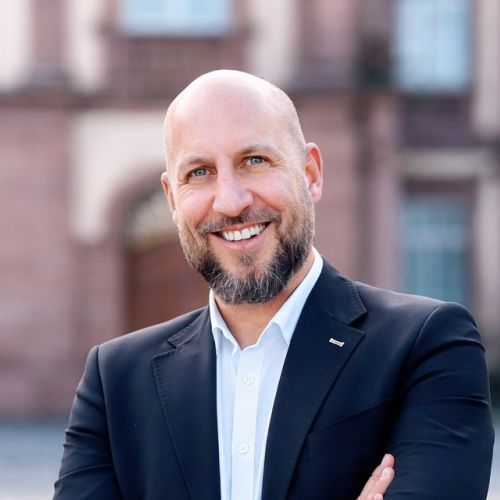 Mann mit Bart und Glatze in Anzug lächelt vor einem historischen Gebäude.