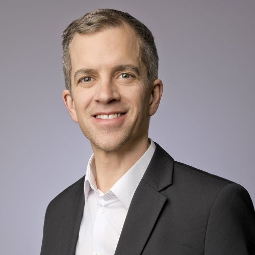 Porträt eines lächelnden Mannes in Anzug mit grauem Hintergrund, der Tobias Bauer repräsentiert.