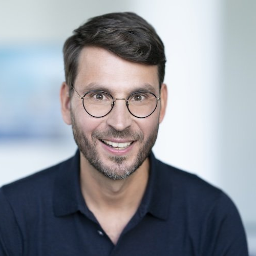 Porträt eines lächelnden Mannes mit Brille und kurzem, dunklem Haar, in einem hellen, unscharfen Hintergrund.