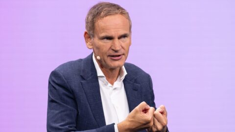 Mann mit Mikrofon spricht auf einer Bühne vor lila Hintergrund.