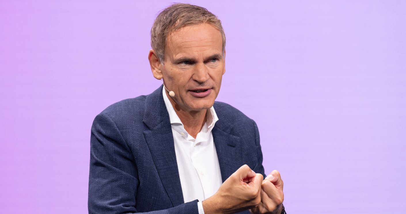 Mann mit Mikrofon spricht auf einer Bühne vor lila Hintergrund.