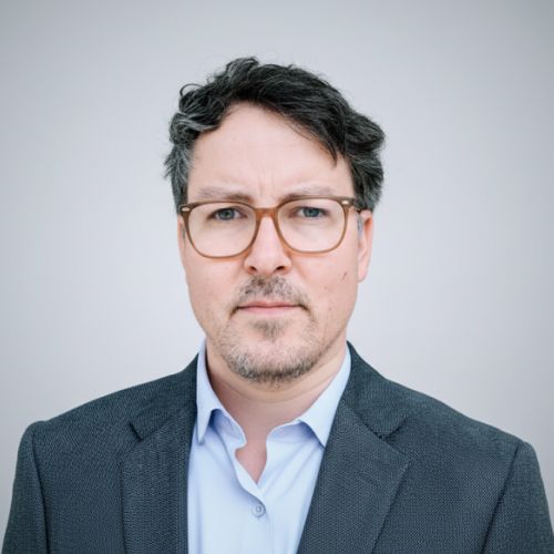 Porträt von Florian Boenigk in einem Anzug mit Brille, neutralem Hintergrund, ernstem Gesichtsausdruck.