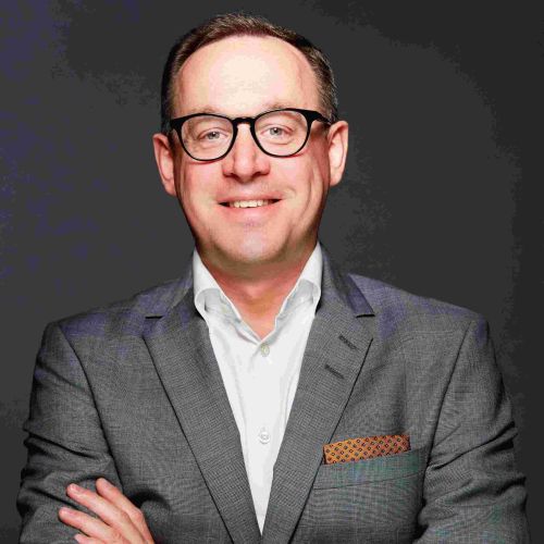 Mann mit Brille und Anzug lächelt, Arme vor der Brust verschränkt, vor dunklem Hintergrund.