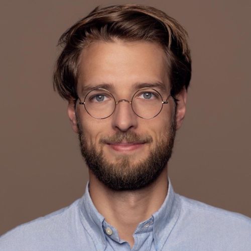 Porträt eines Mannes mit Brille und Bart, lächelnd vor einem neutralen Hintergrund. Er trägt ein hellblaues Hemd.