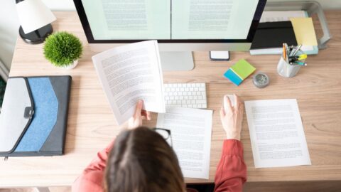 Blick auf einen Schreibtisch mit Computer, an dem eine Frau sitzt und Dokumente prüft.