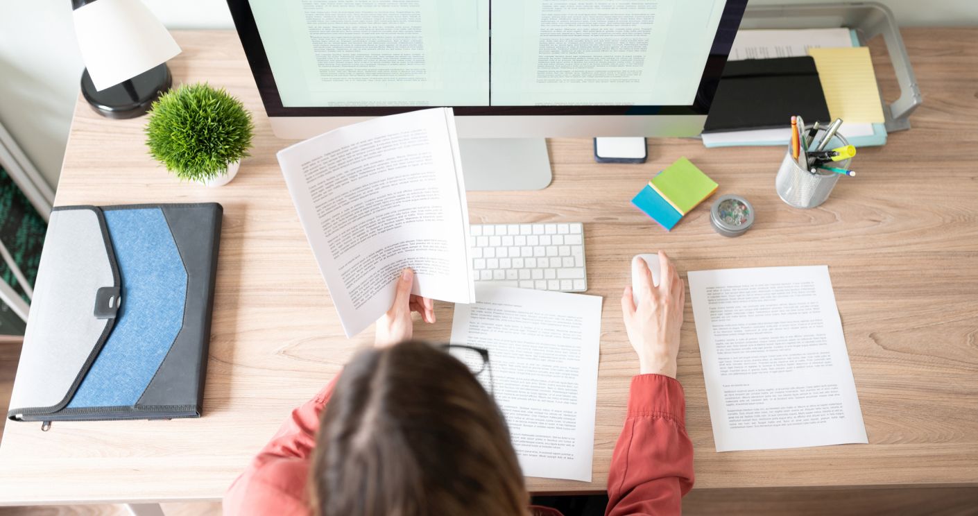 Blick auf einen Schreibtisch mit Computer, an dem eine Frau sitzt und Dokumente prüft.