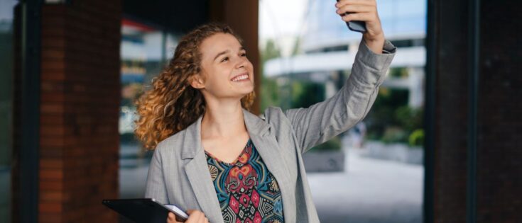 Junge Frau mit lockigem Haar macht ein Selfie, hält einen Laptop und steht vor einem modernen Stadtgebäude.
