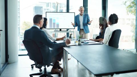 Manager präsentiert Strategien vor einem Team in einem modernen Besprechungsraum mit einem Bildschirm im Hintergrund.
