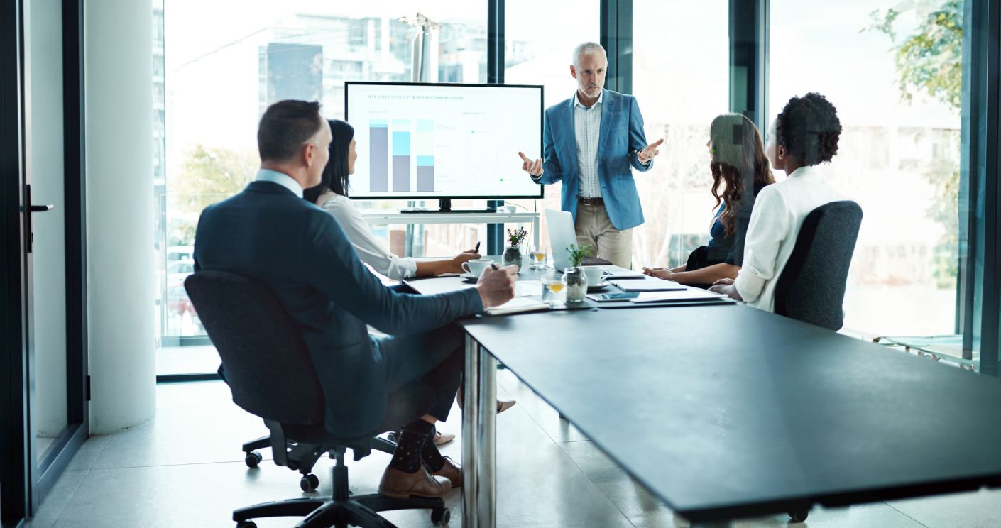 Manager präsentiert Strategien vor einem Team in einem modernen Besprechungsraum mit einem Bildschirm im Hintergrund.