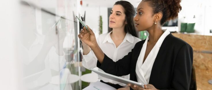 Zwei multikulturelle Frauen analysieren Daten an einem Whiteboard und arbeiten gemeinsam an einem Projekt in einem Büro.