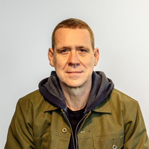 Porträt eines Mannes mit kurzem Haar, in einer Jacke und Kapuzenpullover, vor einem hellen Hintergrund.