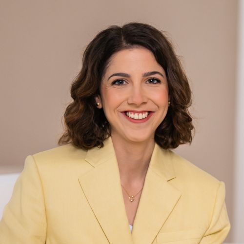 Frau mit lockigem, braunem Haar in einem gelben Blazer lächelt freundlich vor einem neutralen Hintergrund.