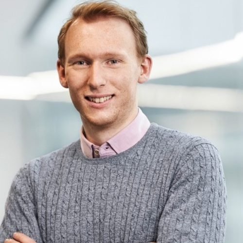 Junger Mann mit blonden Haaren, lächelnd, trägt einen grauen Pullover und ein rosa Hemd, vor einem modernen Hintergrund.