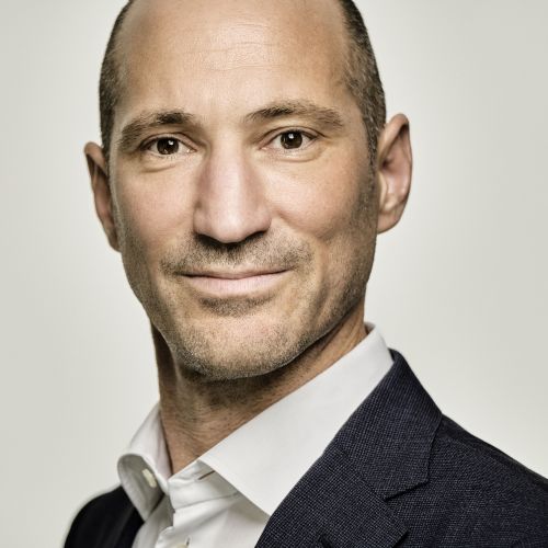 Porträt eines Mannes mit kurzen Haaren, lächelnd, in einem Anzug mit Hemd, neutralem Hintergrund.