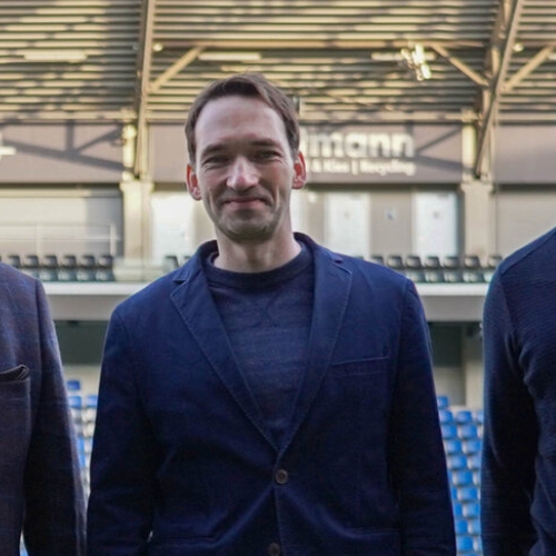 Manuel Holscher steht lächelnd in einem Stadion, umgeben von zwei weiteren Personen, mit leeren Sitzplätzen im Hintergrund.