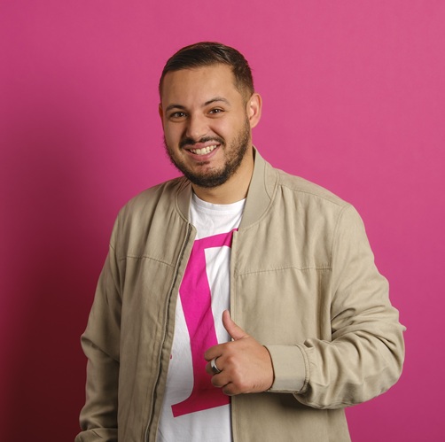 Mann mit Bart und lächelndem Gesicht, trägt eine beige Jacke und ein T-Shirt mit Logo vor pinkem Hintergrund.