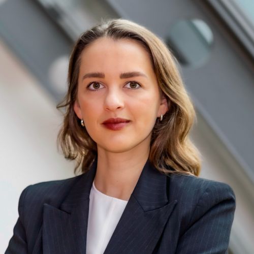 Frau mit langen, lockigen Haaren in einem schwarzen Blazer, lächelt vor einem modernen Hintergrund.