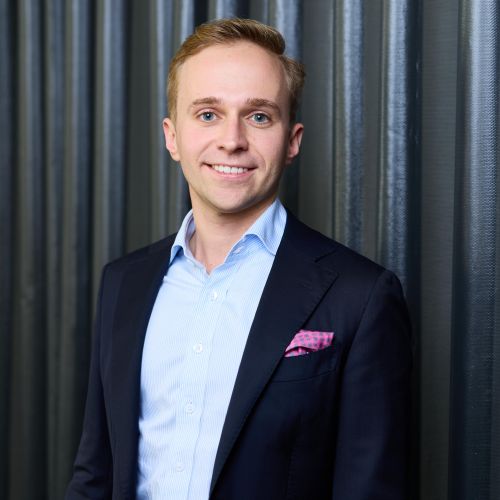 Porträt eines lächelnden Mannes in einem Anzug mit blauer Hemd und pinkem Einstecktuch vor einer strukturierten Wand.