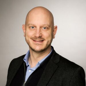 Porträt eines lächelnden Mannes mit Glatze, in einem dunklen Blazer und hellblauer Hemd, vor neutralem Hintergrund.