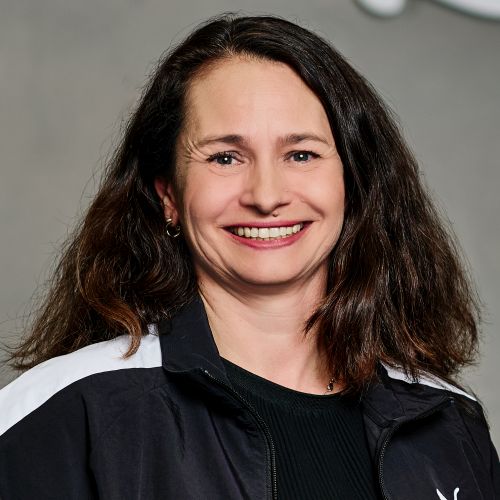 Porträt von Anne Putz mit langen, dunklen Haaren, lächelnd in einer schwarzen Puma-Jacke vor einem grauen Hintergrund.