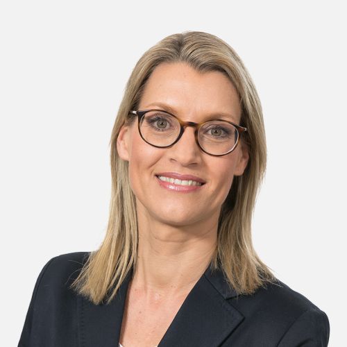 Porträt einer Frau mit blonden Haaren und Brille, die in einem schwarzen Blazer lächelt.