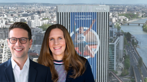 Zwei lächelnde Personen vor einer modernen Stadtlandschaft mit einem großen Werbebanner für Paris 2024.