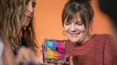 Zwei Frauen halten einen bunten, prismatischen Preis in den Händen, lächelnd vor einem orangefarbenen Hintergrund.