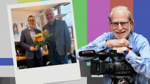 Zwei Männer in einem Büro halten einen Blumenstrauß; einer lächelt, der andere posiert mit einer Kamera.