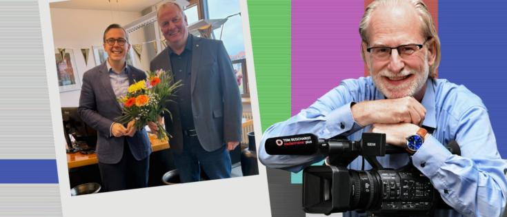 Zwei Männer in einem Büro halten einen Blumenstrauß; einer lächelt, der andere posiert mit einer Kamera.