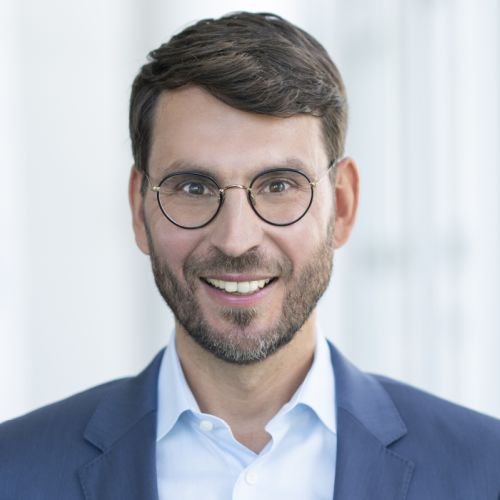 Porträt eines lächelnden Mannes mit Brille, dunklem Haar und Anzug, vor einem hellen, unscharfen Hintergrund.