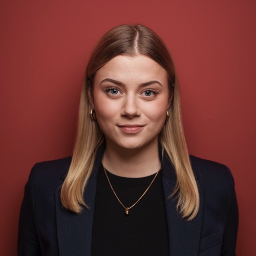 Porträt einer jungen Frau mit blonden Haaren, die vor einem roten Hintergrund steht, in formeller Kleidung.