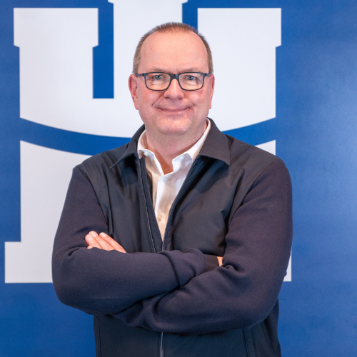 Frank Bleydorn steht mit verschränkten Armen vor einem blauen Hintergrund mit dem Logo des Handball Sport Vereins Hamburg.