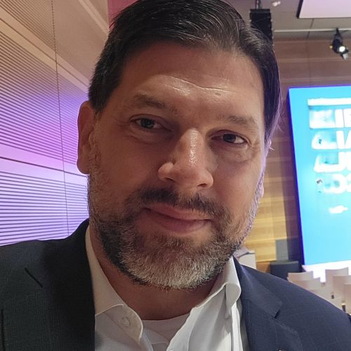 Porträt eines Mannes mit Bart, in einem Anzug, lächelnd vor einem modernen Hintergrund.