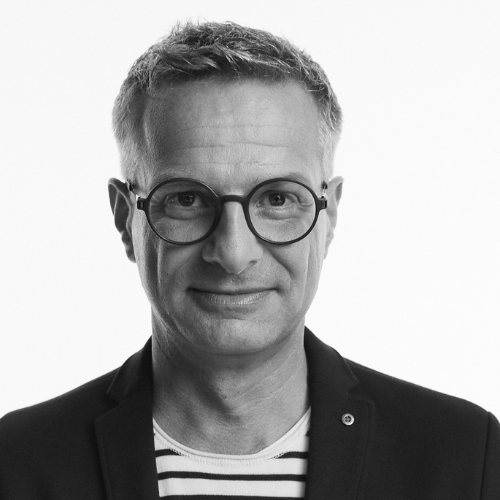 Porträt eines Mannes mit Brille, kurzen grauen Haaren und gestreiftem Shirt, vor einem weißen Hintergrund.