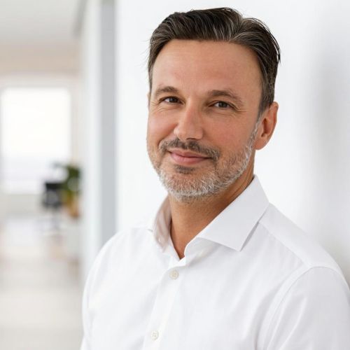 Porträt eines Mannes mit kurzen, dunklen Haaren und Bart, der in einem hellen Raum lächelt. Er trägt ein weißes Hemd.