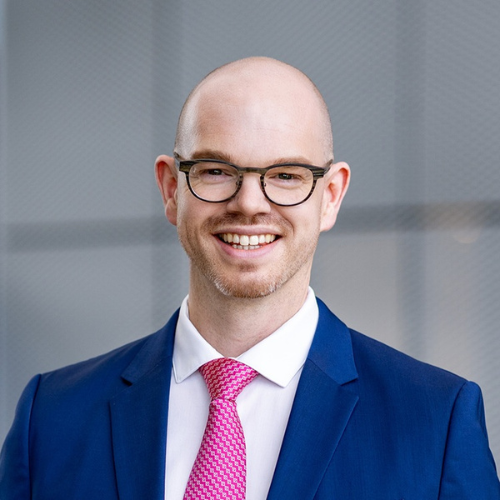 Mann mit Brille und Glatze, trägt einen blauen Anzug und eine pinke Krawatte, lächelt vor einem modernen Hintergrund.