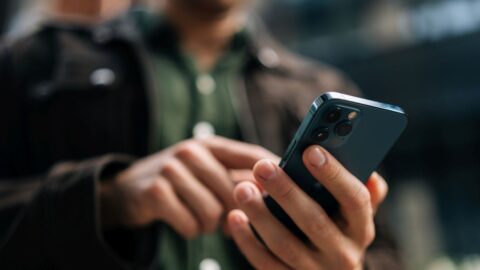 Nahaufnahme von Händen eines Mannes, der ein Smartphone hält und auf einer Stadtstraße das Internet durchsucht.