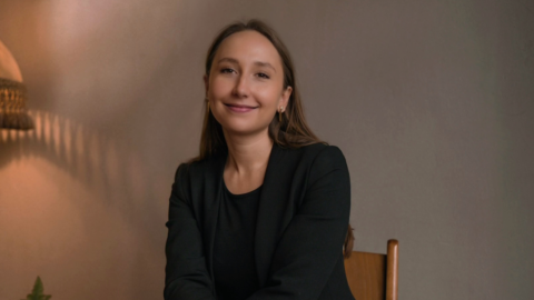 Frau mit langen Haaren in schwarzem Blazer sitzt lächelnd auf einem Stuhl vor einem neutralen Hintergrund.