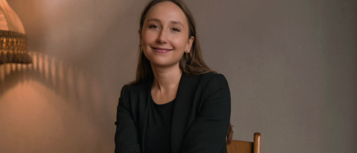 Frau mit langen Haaren in schwarzem Blazer sitzt lächelnd auf einem Stuhl vor einem neutralen Hintergrund.