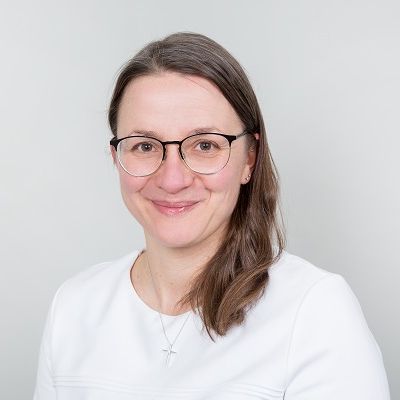 Porträt einer Frau mit Brille und langen Haaren, lächelnd vor einem hellen Hintergrund.