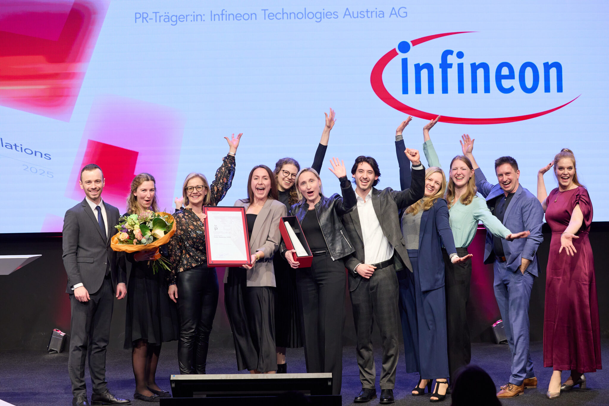 Gruppe von Menschen feiert die Verleihung des PR-Staatspreises an Infineon mit Urkunde und Blumenstrauß.