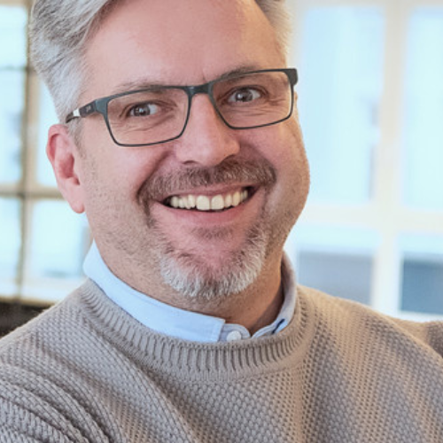 Lächelnder Mann mit Brille und grauem Pullover, Hintergrund mit unscharfen Fenstern.