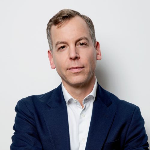 Porträt eines Mannes mit kurzen, hellen Haaren, in einem blauen Blazer und weißem Hemd, vor einem neutralen Hintergrund.