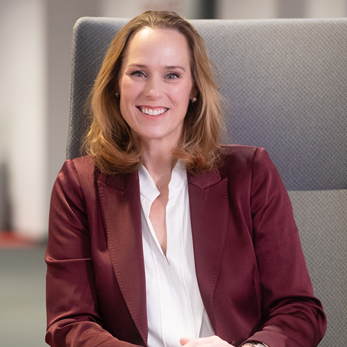 Frau mit schulterlangen, braunen Haaren in einem burgunderfarbenen Blazer, lächelt in einem modernen Büroumfeld.