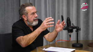 Mann mit Bart spricht leidenschaftlich in ein Mikrofon, Papiere und weiteres Equipment auf dem Tisch.